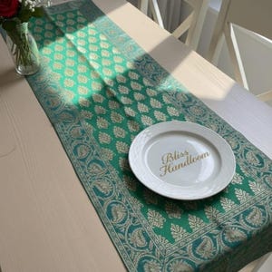 以下が含まれることがあります： 緑と金のフローラル柄のテーブルランナー。白いお皿には「Bliss Handloom」と書かれています。ランナーは繰り返しの花柄のデザインです。左側には赤いバラの花瓶があります。