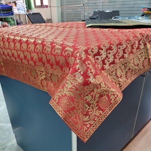 Pode incluir: Uma toalha de mesa vermelha com um padrão de elefantes dourados e uma borda floral. A toalha de mesa está drapeada sobre uma superfície azul, mostrando seu design intrincado e sua cor rica. O tecido parece ser um material tecido.