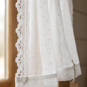 Könnte beinhalten: Ein weißer, transparenter Vorhang mit einem zarten Punktmuster und einer bogenförmigen Spitzenborte an der vertikalen Kante. Der Stoff ist leicht und halbtransparent, mit einem schlichten weißen Saum am unteren Ende und einem kleinen braunen Etikett.