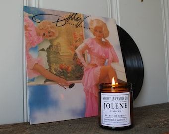 Puede incluir: Una vela de vidrio marrón con una etiqueta blanca que dice "Nashville Candle Co. Jolene Hibiscus & Breath of Spring Hand Poured in Nashville, TN NashvilleCandleCompany.com". La vela está encendida y la llama es visible. La vela está sentada sobre una superficie de madera frente a un disco de vinilo con una imagen de Dolly Parton en la portada.