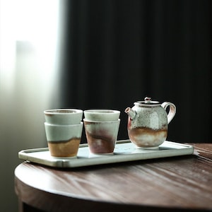 May include: A set of four small ceramic teacups and a teapot with a speckled glaze in shades of brown, white, and green. The cups and teapot are arranged on a rectangular tray.