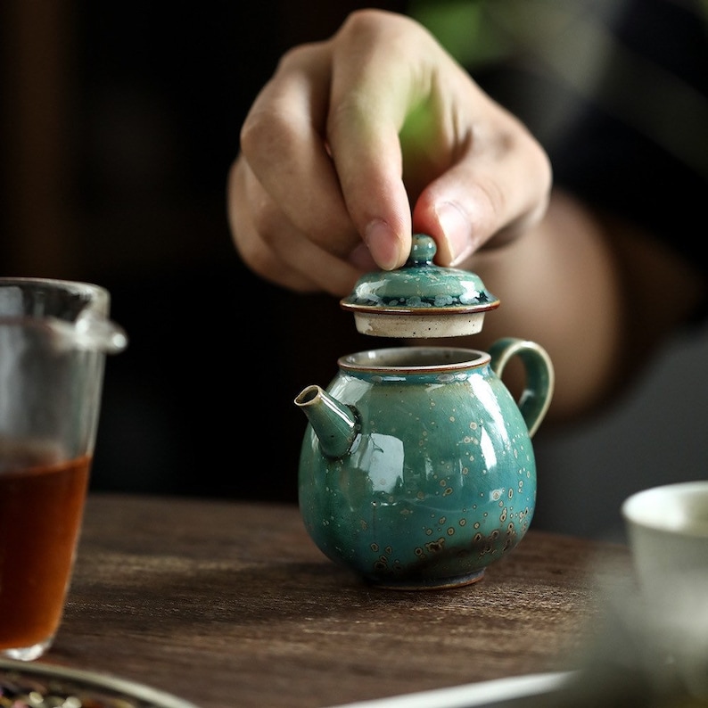 May include: A small, round teal ceramic teapot with a matching lid. The teapot has a spout and a handle. The lid is being lifted, revealing the interior. The teapot has a speckled glaze and a brown rim. Tea set.