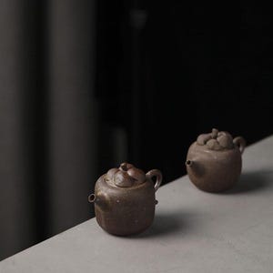 Può includere: Due piccole teiere in ceramica marrone con superfici testurizzate e coperchi a forma di frutta. Le teiere sono su una superficie grigio chiaro.