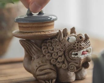 May include: A ceramic tea set featuring a brown and white guardian lion figurine with a removable lid. The lid is a light blue ceramic disc with a brown rim.