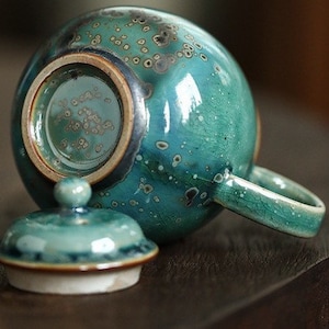 May include: A teal ceramic teapot with a matching lid. The teapot has a round body, a handle, and a spout. The lid has a small knob. The teapot has a speckled pattern and a glossy finish.