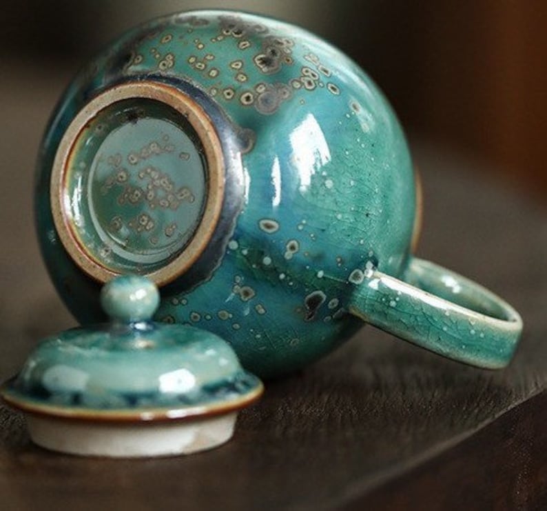 May include: A teal ceramic teapot with a matching lid. The teapot has a round body, a handle, and a spout. The lid has a small knob. The teapot has a speckled pattern and a glossy finish.