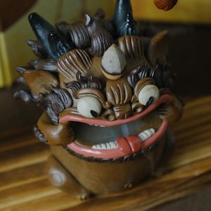 May include: A detailed wooden sculpture of a mythical creature with a wide open mouth, visible teeth, and a pink tongue. The sculpture has large eyes, horns, and detailed fur. The creature is brown with black accents and sits on a wooden surface.