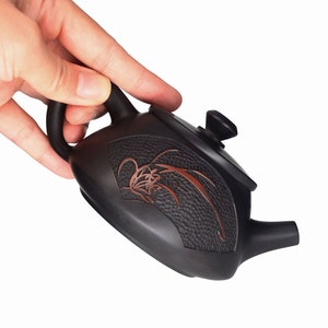 May include: A black teapot with a brown floral design and a textured surface. The teapot has a lid with a knob on top and a spout on the side.