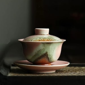 Może przedstawiać: Ceramiczny kubek do herbaty w kolorze różowym i zielonym z pokrywką i pasującym spodkiem. Kubek do herbaty ma unikalny, marmurowy wzór.