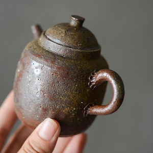 May include: A small, brown ceramic teapot with a textured surface and a rounded handle. The teapot has a lid with a small knob on top.