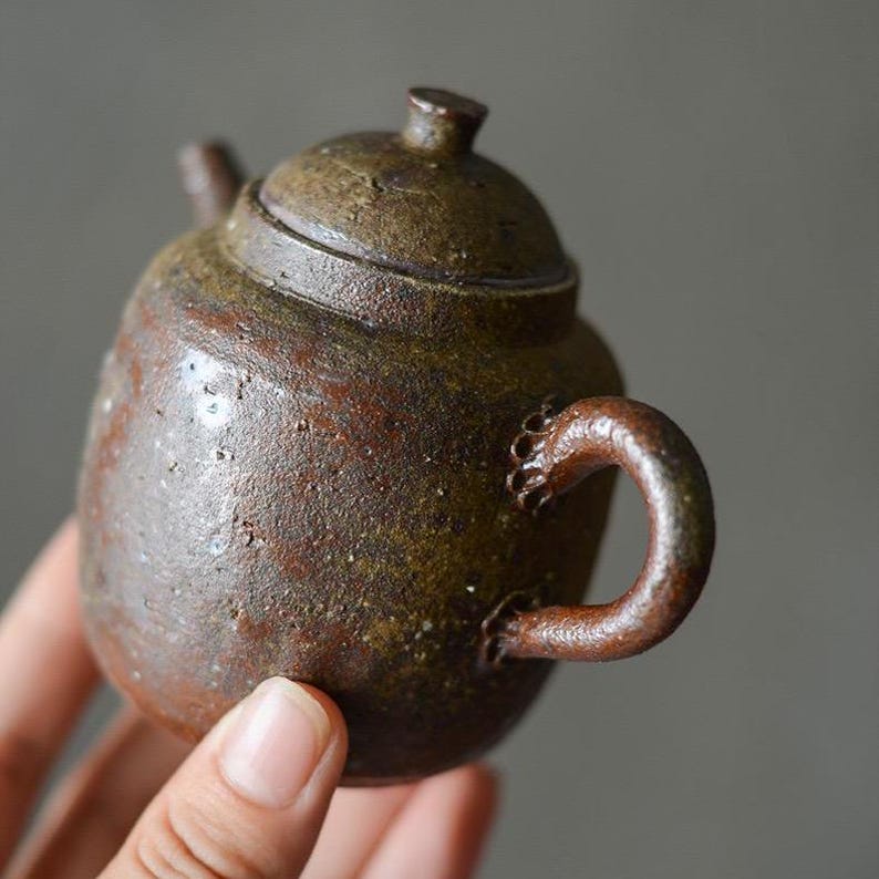 May include: A small, brown ceramic teapot with a textured surface and a rounded handle. The teapot has a lid with a small knob on top.