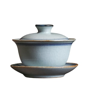 Peut inclure: Une théière en céramique blanche avec un couvercle et une soucoupe assortie. La théière a un bord bleu et brun et un glaçage craquelé.