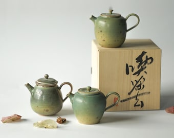 Könnte beinhalten: Drei grüne Teekannen aus Keramik mit gesprenkeltem Überzug. Die Teekannen stehen auf einer Holzkiste mit japanischen Schriftzeichen.