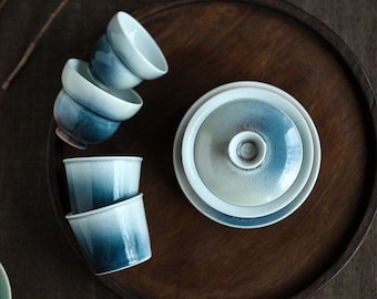 Könnte beinhalten: Ein Set aus Keramik-Teetassen und einer Teekanne mit Deckel und blauem Farbverlauf. Die Tassen sind auf einem dunklen Holztablett gestapelt. Das Teeset hat ein minimalistisches Design und eine moderne Ästhetik.