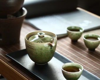 May include: A ceramic tea set featuring a lidded gaiwan and three small matching cups, all finished in a mottled green and cream glaze. The pieces are arranged on a rectangular black tray, showcasing a smooth, rounded design with a small spout.