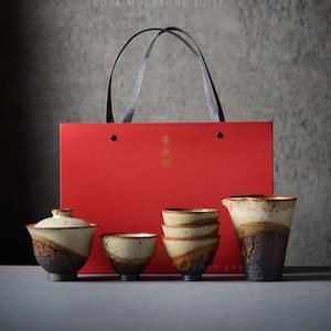 Op de afbeelding: Een theeset met een rode geschenkzak. De set bevat een theepot met deksel, kleine kommen en een kan. De keramische stukken hebben een gespikkelde beige en bruine glazuur. De geschenkzak heeft zwarte handvatten en Chinese karakters.