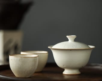 May include: White ceramic tea set with a teapot and two cups on a wooden tray. The teapot has a lid and a small spout. The cups are small and round. The tray is dark brown and has a smooth surface.