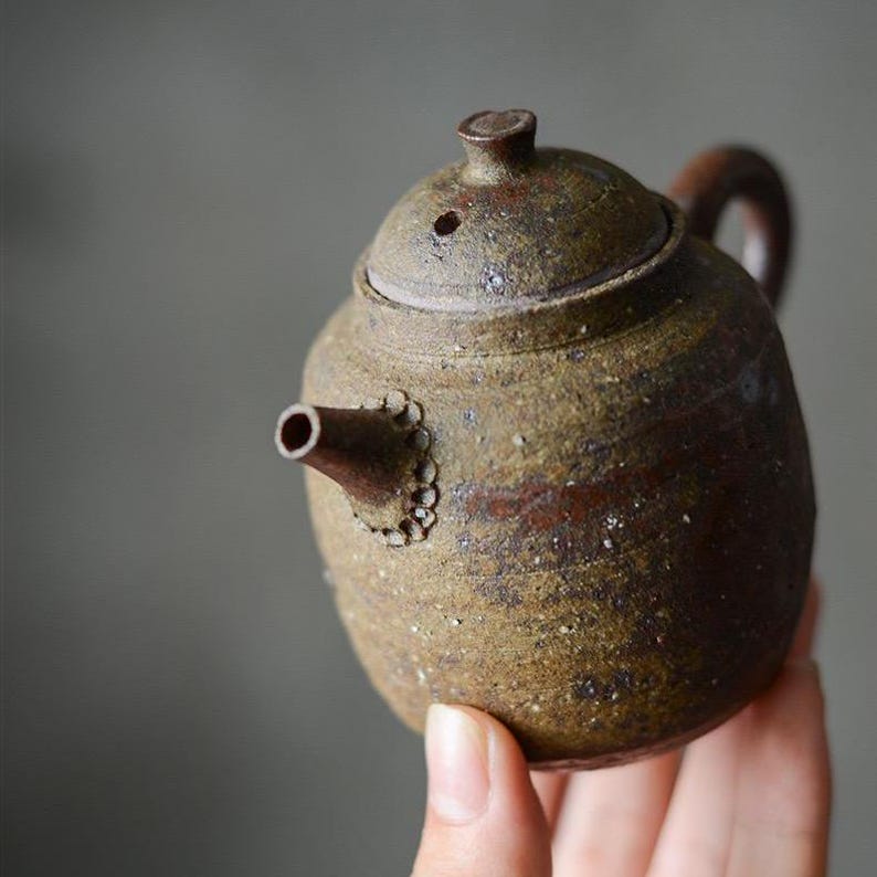 May include: A small, brown, speckled teapot with a lid and a spout. The teapot has a decorative band of small circles around the spout.
