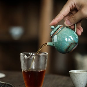 May include: A small, turquoise ceramic teapot with a brown rim is pouring tea into a clear glass. The teapot has a handle and a lid. The tea is dark brown. A small white ceramic cup is in the background.