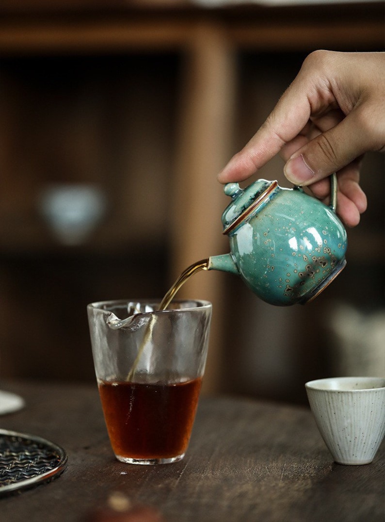 May include: A small, turquoise ceramic teapot with a brown rim is pouring tea into a clear glass. The teapot has a handle and a lid. The tea is dark brown. A small white ceramic cup is in the background.
