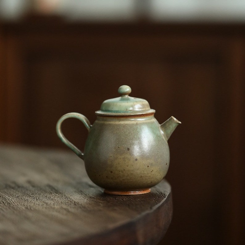 May include: A small, round ceramic teapot with a lid and handle. The teapot is primarily olive green with brown accents and a small spout. The lid has a small knob on top. The teapot is sitting on a dark wooden surface.
