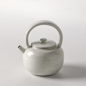 Op de afbeelding: Een witte keramische theepot met een afgeronde body, een gebogen handvat en een kleine tuit. Het deksel heeft een kleine, ronde knop. De theepot is licht, gebroken wit van kleur en staat op een wit oppervlak.