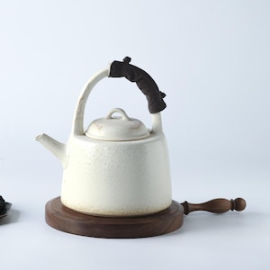 May include: Cream-colored ceramic teapot with a curved handle and a dark brown padded grip. The teapot sits on a round, dark wooden base with a matching handle. The lid has a small, rounded handle.