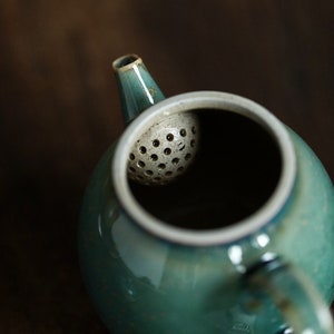 May include: A close-up of a teal ceramic teapot with a white rim and spout. The teapot has a built-in, perforated tea infuser. The background is a blurred, dark brown.