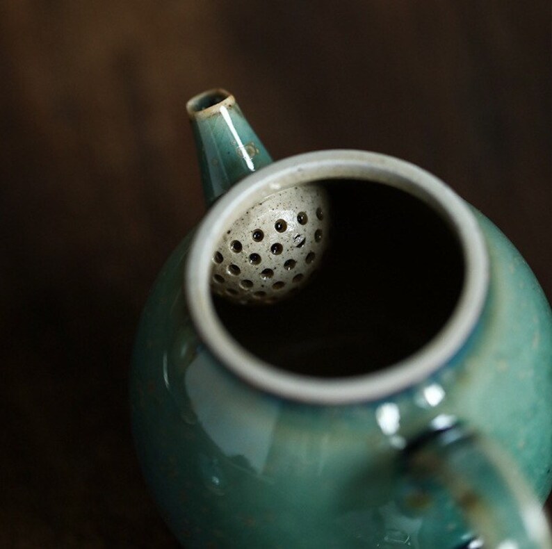 May include: A close-up of a teal ceramic teapot with a white rim and spout. The teapot has a built-in, perforated tea infuser. The background is a blurred, dark brown.