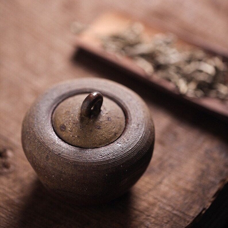 May include: A small, round, brown ceramic jar with a lid and a handle. The jar is sitting on a wooden surface.