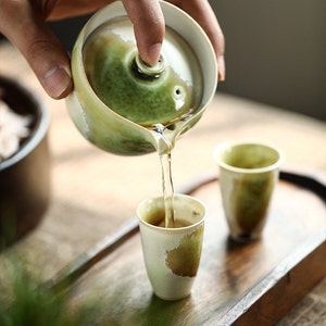 May include: A green and white ceramic teapot pouring tea into a matching teacup. The teapot has a lid and a spout. The teacup is sitting on a wooden tray.