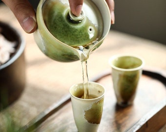 May include: A green and white ceramic teapot pouring tea into a matching teacup. The teapot has a lid and a spout. The teacup is sitting on a wooden tray.