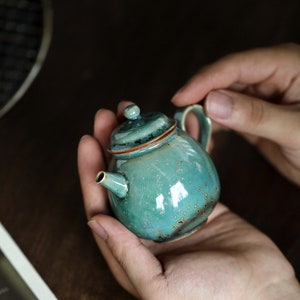 May include: A small, turquoise ceramic teapot with a rounded body, a lid, and a handle. The teapot has a glossy finish with brown accents and is held in open hands. The teapot is a decorative item.