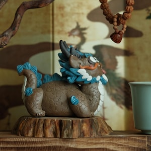 Puede incluir: Figura de dragón de cerámica marrón con detalles azules y blancos, sobre una base de madera. A la derecha hay una taza de té azul claro, con una rama decorativa de madera y un adorno de cuentas colgante en el fondo.