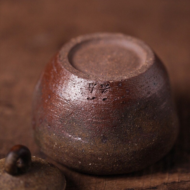 May include: A brown ceramic teacup with a rough, textured surface. The bottom of the cup has a small, dark inscription.