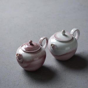 Peut inclure: Deux théières en céramique avec un glaçage blanc et un dégradé de rose et de violet. Les théières ont un corps arrondi et un petit bec.