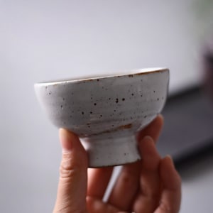 May include: A small, off-white ceramic bowl with a speckled surface and a slightly flared rim. The bowl has a short, pedestal base and a rustic, handmade appearance. The rim has a brown accent.