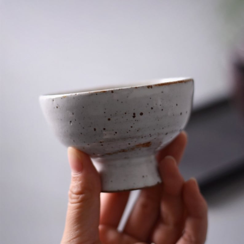 May include: A small, off-white ceramic bowl with a speckled surface and a slightly flared rim. The bowl has a short, pedestal base and a rustic, handmade appearance. The rim has a brown accent.