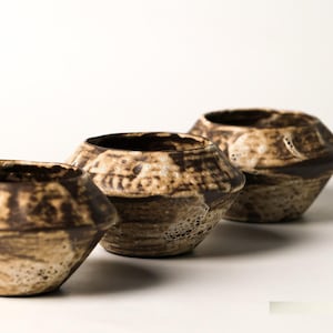 May include: Three ceramic bowls arranged in a row, each featuring a wide, angular midsection and a tapered base. The bowls have a textured, mottled surface with a colour palette of dark brown and cream, showing a rustic, uneven glaze finish.