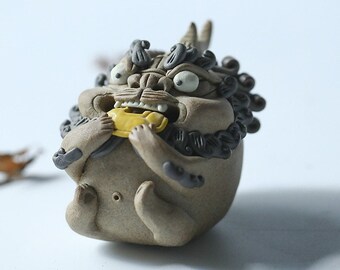 May include: A ceramic figurine of a mythical creature with a large head, wide eyes, and a long, flowing mane. The creature is holding a golden object in its mouth. The figurine is painted in shades of brown and black.