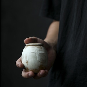 以下が含まれることがあります： 蓋付きの小さな手作りの陶器の壺。壺は丸みを帯びた形で、茶色のアクセントが付いた、テクスチャ加工されたオフホワイトの釉薬が施されています。蓋は同じ色です。壺は、暗い背景に対して人の手で持たれています。