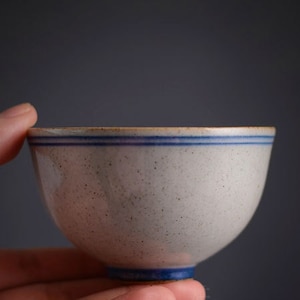 May include: A small, off-white ceramic bowl with speckled texture and two thin blue stripes near the rim. The base of the bowl is also blue. The bowl is held in a hand against a dark gray background.