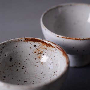 May include: Two white ceramic bowls with a speckled pattern and a rustic, brown rim. The bowls have a simple, minimalist design, suitable for serving food or as decorative items. The bowls are set against a neutral background.