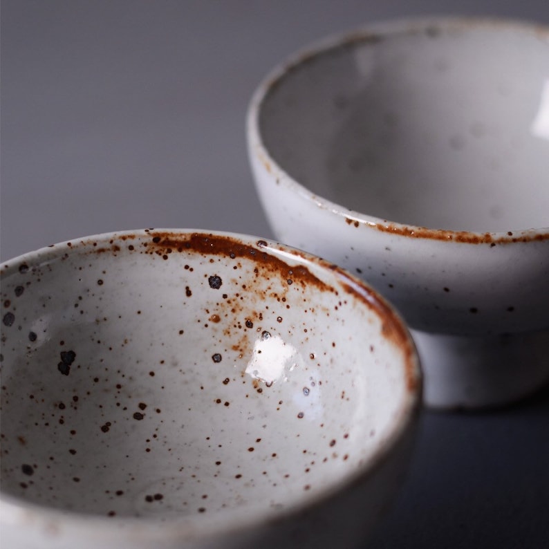 May include: Two white ceramic bowls with a speckled pattern and a rustic, brown rim. The bowls have a simple, minimalist design, suitable for serving food or as decorative items. The bowls are set against a neutral background.