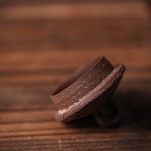 May include: A close-up of a brown ceramic lid with a rough texture. The lid is slightly tilted and appears to be made of clay.