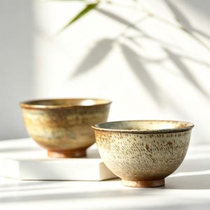 May include: Two ceramic bowls with a rounded shape and a footed base. The front bowl features a textured, cream-colored exterior with brown speckled patterns, while the bowl behind it displays a smooth, blended brown and tan glaze finish.
