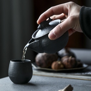 Könnte beinhalten: Eine schwarze Teekanne aus Keramik mit einem fischförmigen Deckel, die Tee in eine schwarze Keramiktasse gießt. Die Teekanne hat eine strukturierte Oberfläche und einen kleinen Ausguss. Die Tasse ist rund und hat einen breiten Boden.