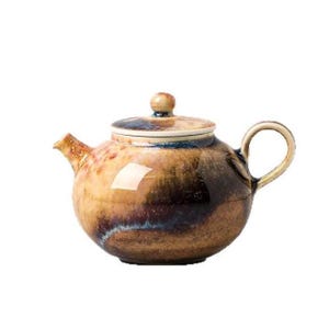 Puede incluir: Una tetera de cerámica con cuerpo redondeado y tapa a juego. La superficie presenta un esmalte moteado con tonos mezclados de marrón tierra, beige y azul profundo, creando una apariencia texturizada en todo el exterior del recipiente.