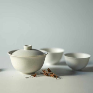 Può includere: Una teiera in ceramica bianca con coperchio e tre tazze abbinate. La teiera e le tazze sono su una superficie bianca con foglie secche sparse intorno.