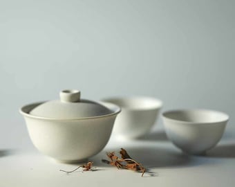 May include: A white ceramic teapot with a lid and three matching teacups. The teapot and cups are sitting on a white surface with dried leaves scattered around them.